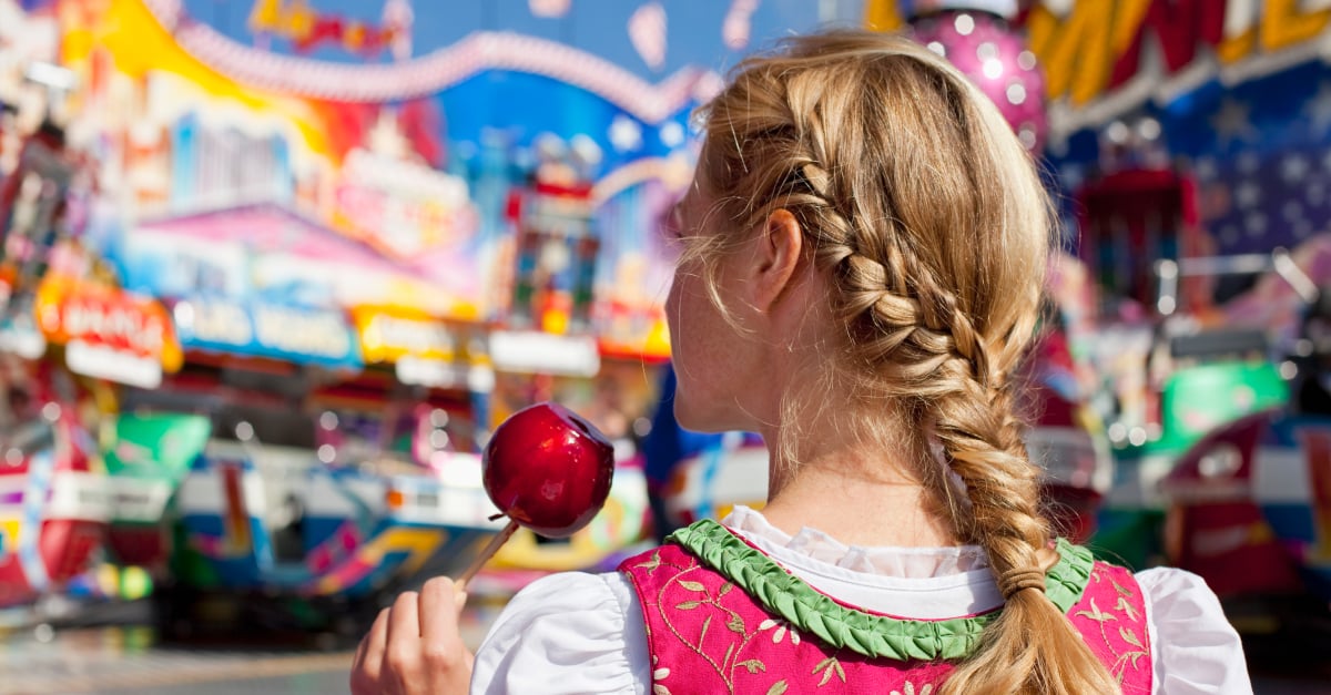 Ein junge Frau besucht ein Volksfest. Sie hält einen karamellisierten 'Apfel in der Hand. Man sieht sie nur von hinten - sie hat ihre blonden Haare zu einem Zopfkranz gebunden.