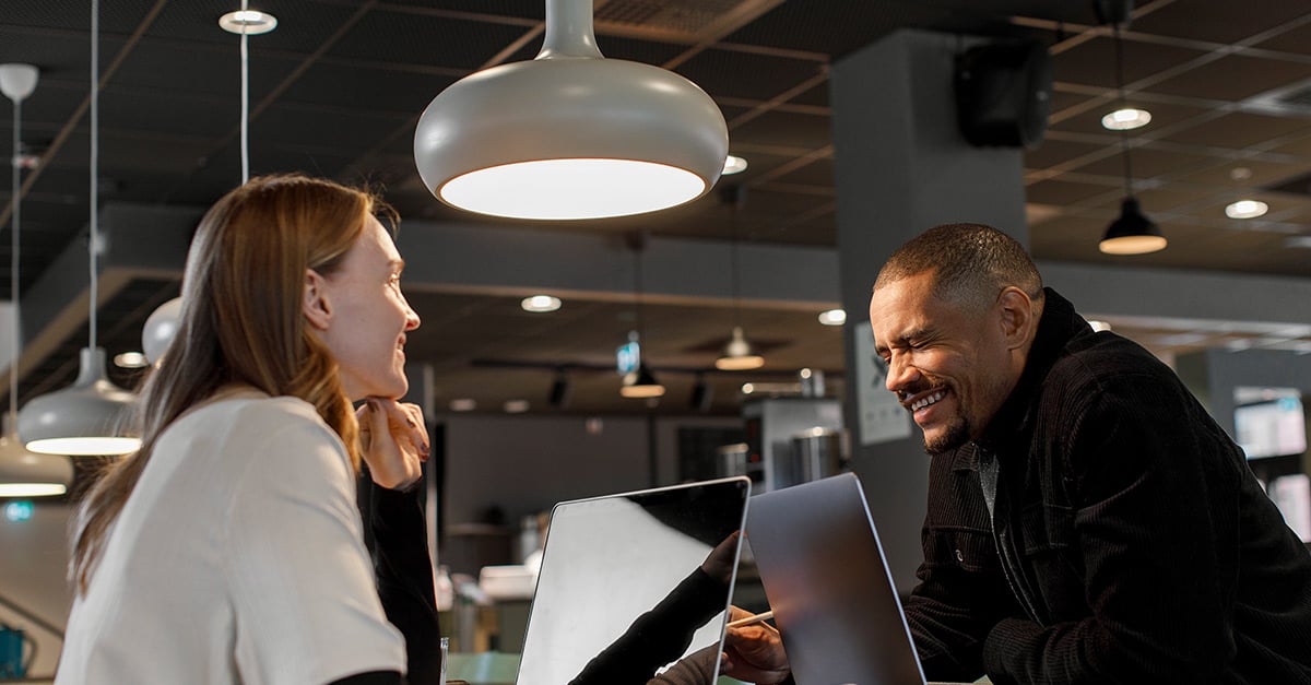 Eine junge Frau und ein junger Mann sitzen in einer Bar. Jede:r hat seinen Laptop vor sich. Es hat den Anschein, dass sie flirten.